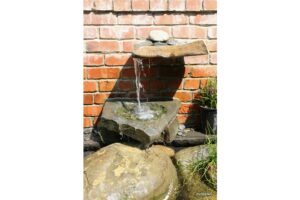 Miniature water feature against brick wall with water plants in pots nearby.