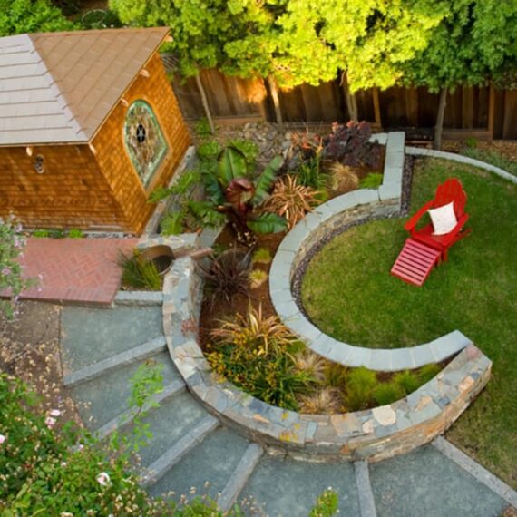 Aerial shot of curved backyard design with plants, stone steps, lawn, chair and guest house.